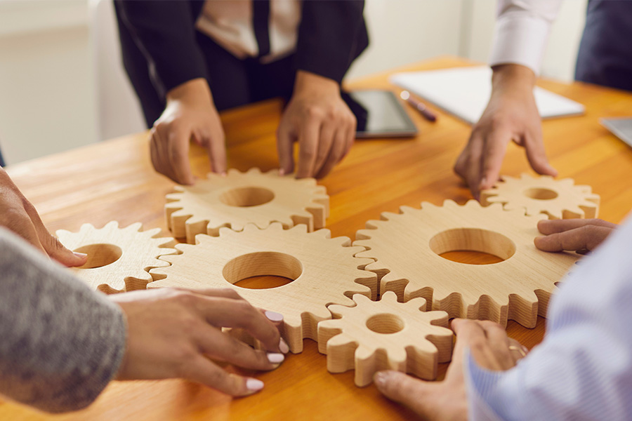 Mehrere Hände von Geschäftsleuten greifen nach ineinandergreifenden Holzzahnrädern auf einem Holztisch. Das Bild symbolisiert Teamarbeit, Zusammenarbeit und effiziente Prozesse in einem Unternehmen oder einer Organisation.